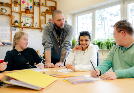 Ein Mann steht an einem Tisch, an dem drei Menschen mit Behinderung sitzen.