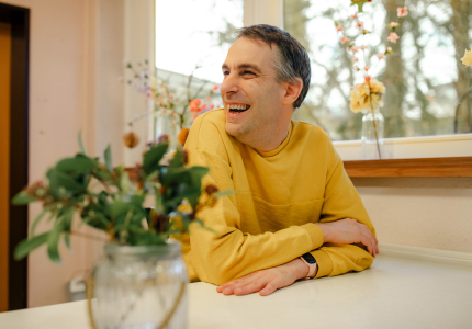 Ein Mann sitzt an einem Tisch und lächelt. Im Vordergrund steht eine Vase